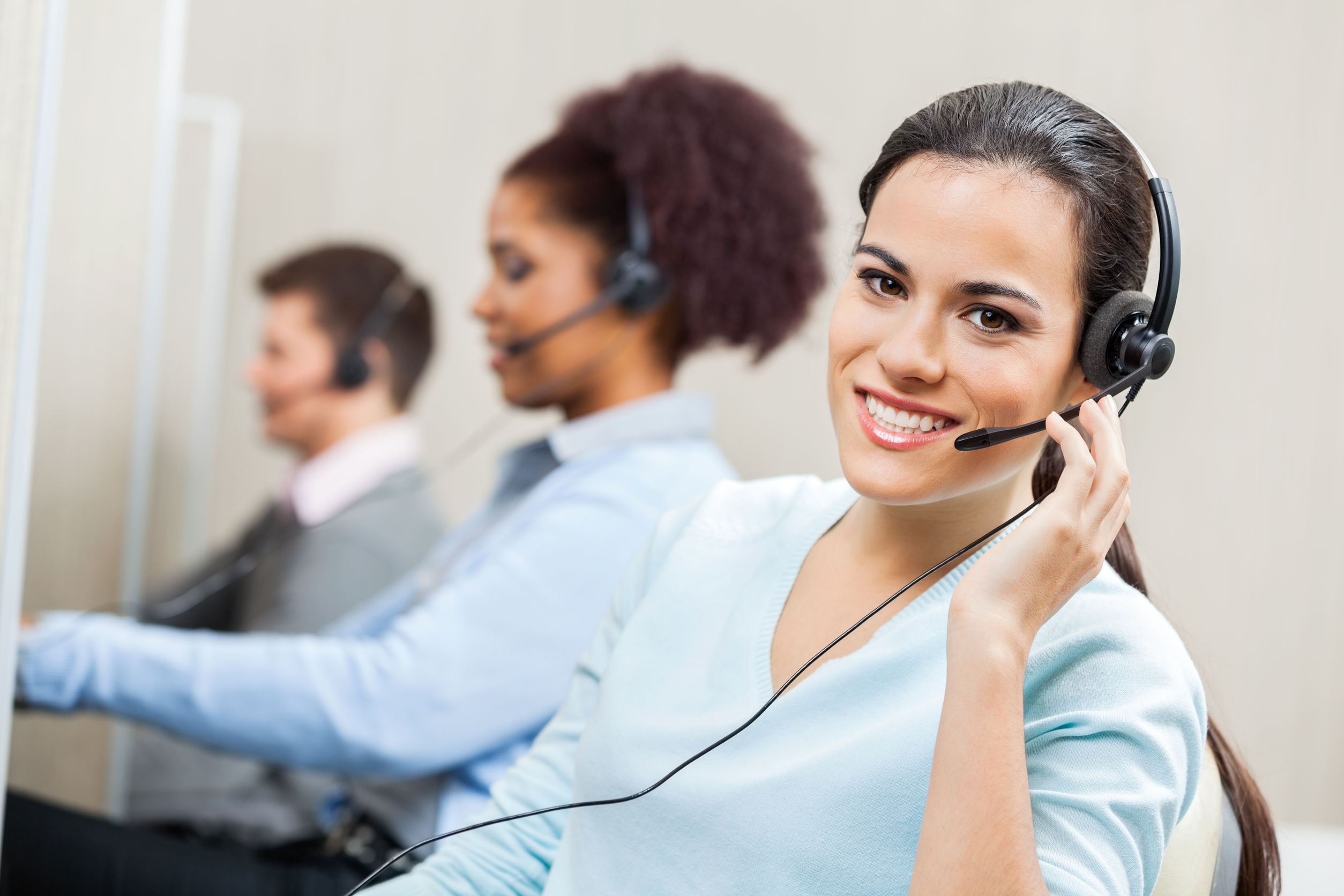 Portrait of smiling female customer service agent wearing headset with colleagues working in background at office